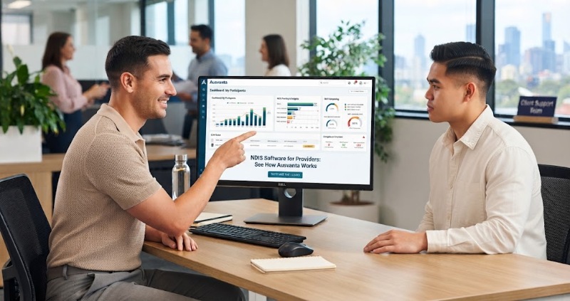Two men collaborate at a desk in a sunlit office with a city view. The man on the left points to a Dell monitor displaying an 'Ausvanta' NDIS software dashboard, while the man on the right looks on.