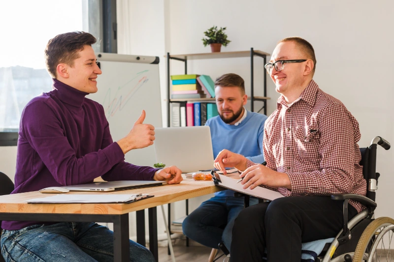 A man in a wheelchair with glasses and a plaid shirt holds a clipboard, smiling while discussing documents with a seated colleague who gives a thumbs-up.